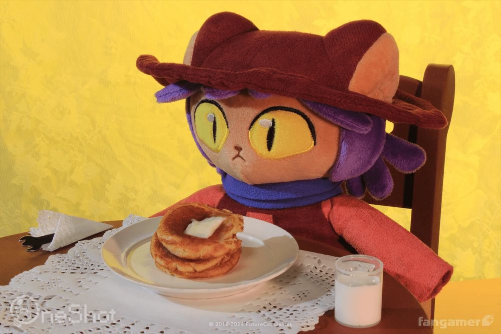 A photo of the Niko plush at a small dinner table. A plate full of pancakes and a glass of milk are on the table. 