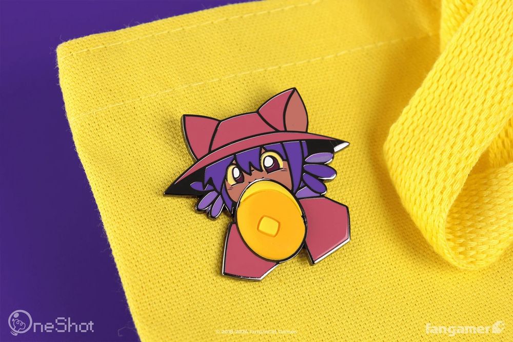A photo of a OneShot pin on a bright yellow tote bag, against a purple background. A hard-enamel Niko is taking a bite out of a distorted, PVC pancake. 