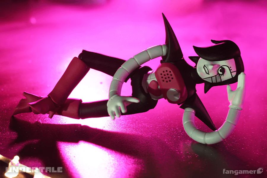 A photograph of the posable Mettaton EX figurine off its base, shot against a hazy fuchsia background. The figurine is lying on its side, posed as though its head is resting on its arm, one leg crossed high over the other.
