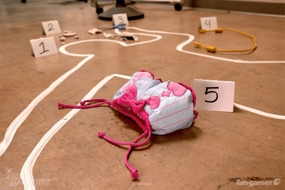 A photo of our new Trucy's Magic Bag drawstring bag photographed in a set designed to look like a crime scene, with numbers and a faux taped-off body. The bag is blue with pink pinstripes, with hearts and a bow on the front and a pink enclosure. 