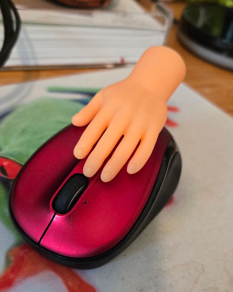 Tiny (finger puppet) hand sitting on a red computer mouse.  Looks like a disembodied hand using a mouse.