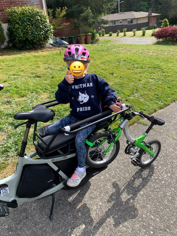 A hangry kid gives a thumbs up while sitting on the back of an e-cargo bike. Her bike is strapped to the back of the e-cargo bike with one wheel trailing on the ground.