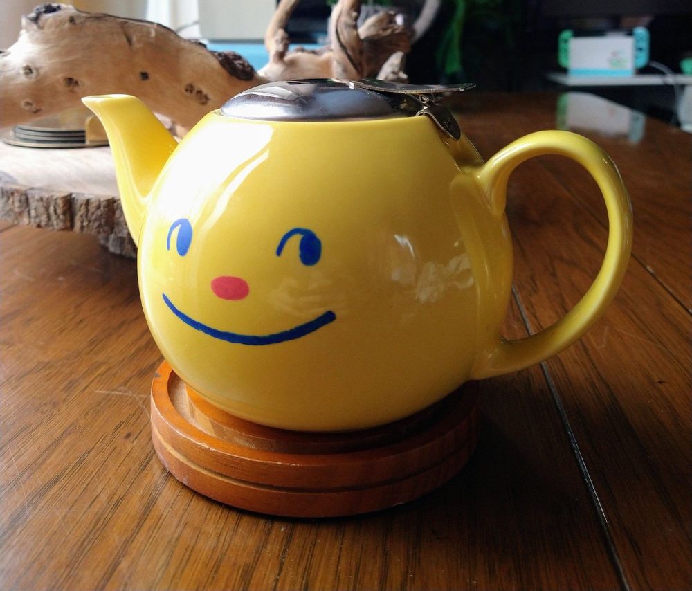 a yellow teapot with a smiley face in blue with a red nose painted on it.