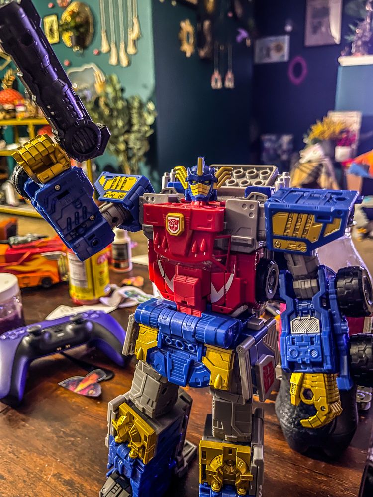 Transformers Legacy Evolution Armada Optimus Prime holding a gun and standing on a cluttered coffee table.