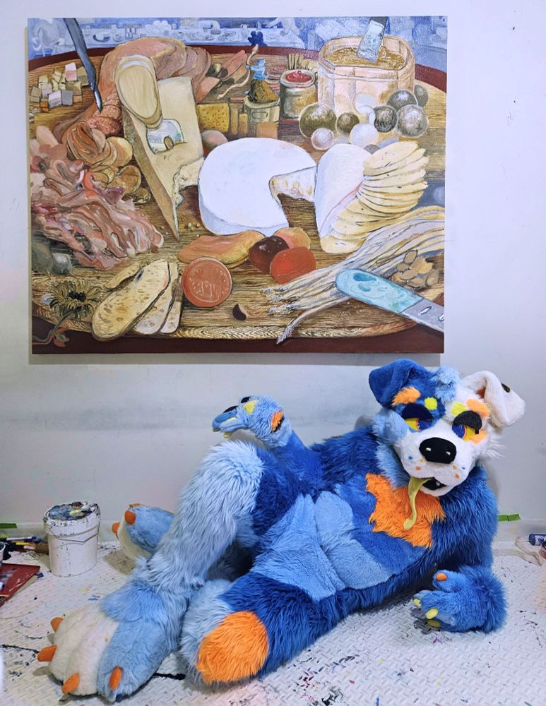 A photo of an artist in their studio with their painting. The artist is laying on the floor, dressed in a custom-tailored blue and orange dog "fursuit", gesturing to the painting above. The painting is 4 x 5 feet in diameter, and depicts a half-eaten charcuterie board with a miniature version of the blue dog moving things around on the board.