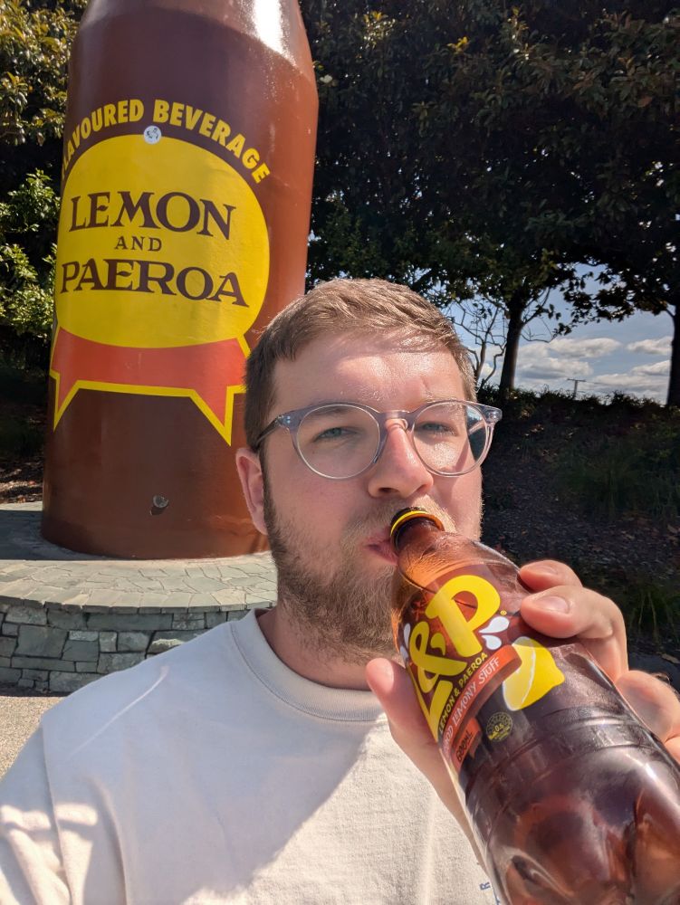 Selfie of Alistair drinking a bottle of L&P — the national drink of New Zealand — in front of a giant concrete L&P bottle