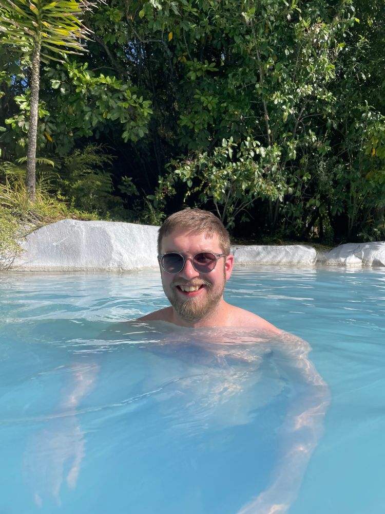 Alistair hanging out in a hot spring with very blue water and lots of foliage in the background. He's got his shades on, is grinning, and looks daft