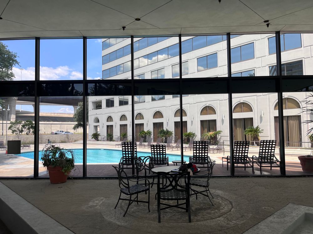 A view through large glass windows showcases an outdoor swimming pool surrounded by lounge chairs and potted plants. The scene includes a modern building with multiple windows and a clear blue sky above.