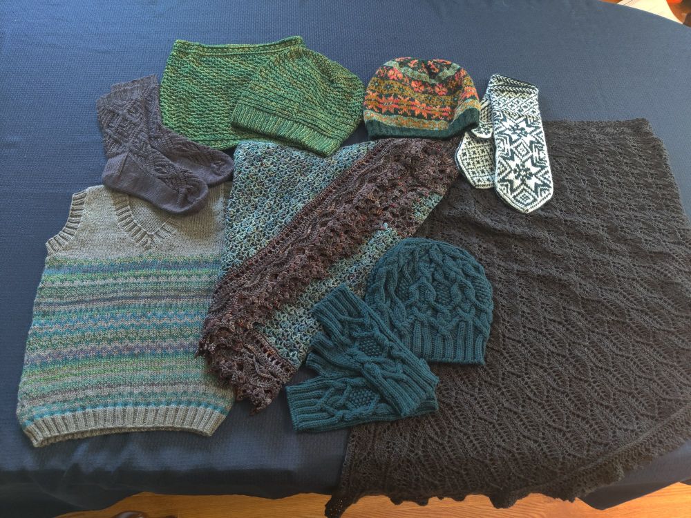 A selection of hand knitted and crocheted items laid out on a blue table cloth. Hats, mitts, socks, shawls, and a child's sleeveless pullover. 