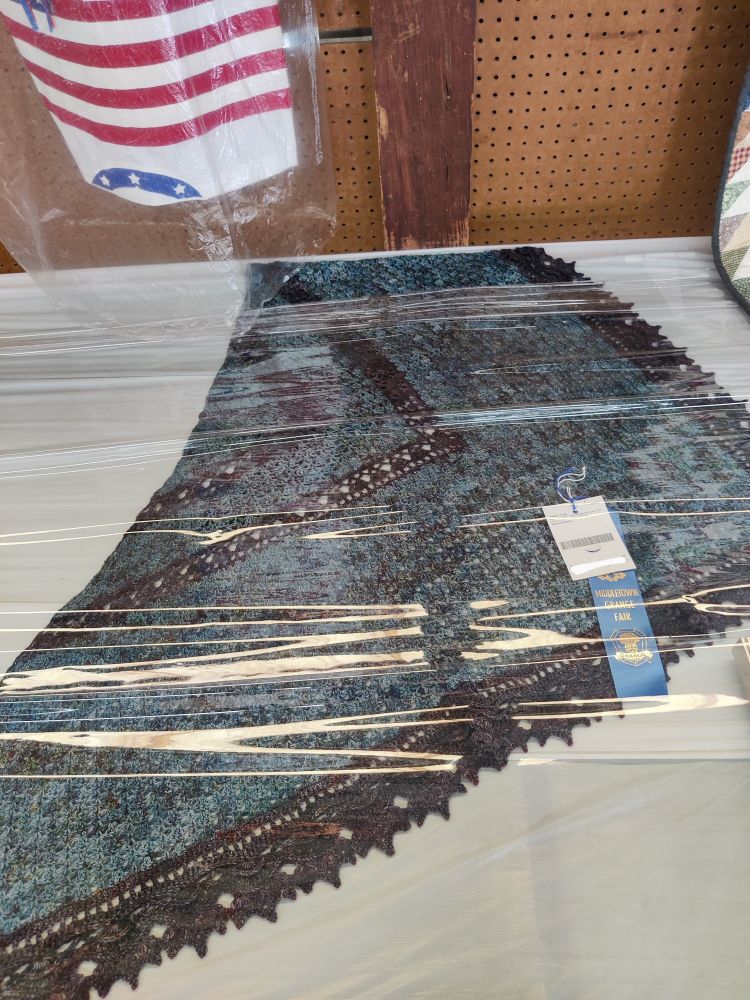 The crochet shawl on full display underneath protective plastic on the Best in Show consideration table. Pattern is Emerald Isle shawl in two colors of Lambstrings Yarn Tralala sock yarn.