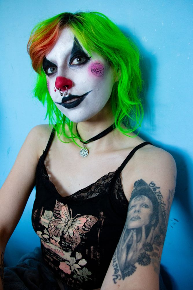 Jinx Aesthel leaning against a blue wall wearing clown makeup, a black choker necklace, black tanktop, and sweatpants 