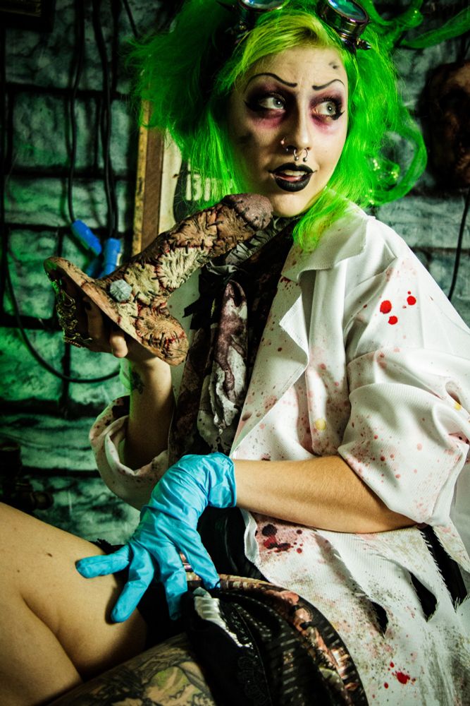 Jinx Aesthel dressed as a mad scientist with fluffy green hair, red eyebags, a blood splattered labcoat and blouse, wearing a glove on one hand and holding the Monstercocks "Frankencock" dildo in the other hand