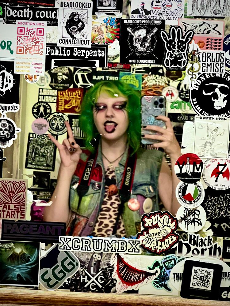 Jinx Aesthel taking a selfie at Cherry Street Station (metal venue in Wallingford, CT) 

the mirror and wall behind Jinx are covered in stickers from various bands, artists, and small businesses

Jinx is holding their phone wearing green hair, gothic makeup, a denim battlevest covered in pins and patches. Under the vest theyre wearing a leopard dress and around their neck is their canon camera
