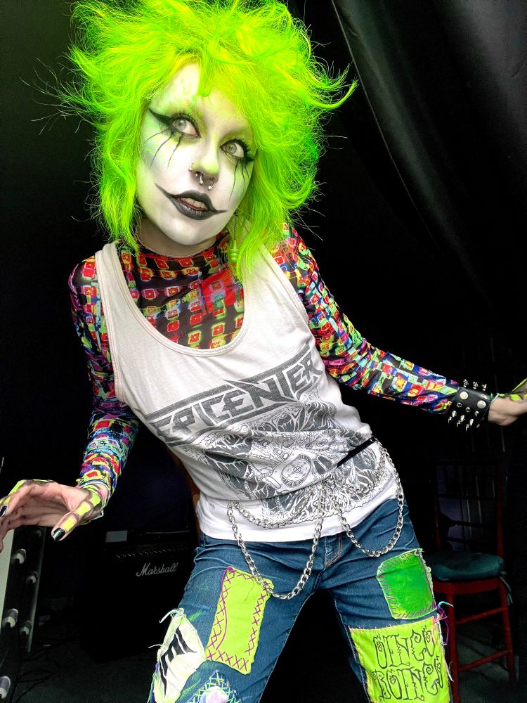 Jinx Aesthel posing with goth makeup, a neon undershirt, Epicenter tank top, and patchwork pants.
