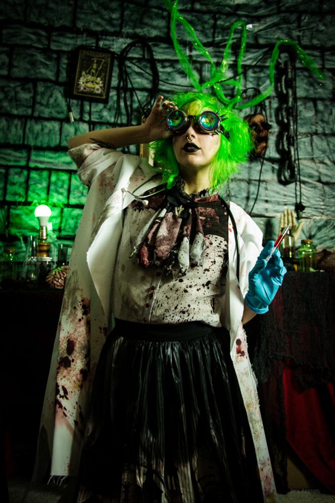 Jinx Aesthel modeling a photoshoot dressed as a mad scientist, holding a syringe with a gloved hand, wearing a lab coat and a bloody blouse and skirt