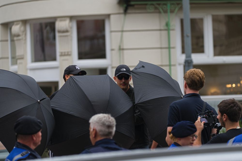 IB-Aktivisten schirmen Presse-Vertreter*innen mit schwarzen Regenschirmen auf dem Lueger-Platz ab. Drei Polizist*innen des LPD-Dialogteams stehen davor und beobachten das Geschehen.