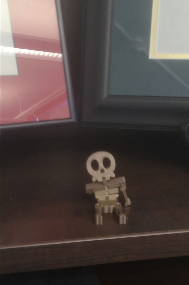 A tiny bronze plastic skeleton sits on a shelf in front of two frames. He belongs there. 