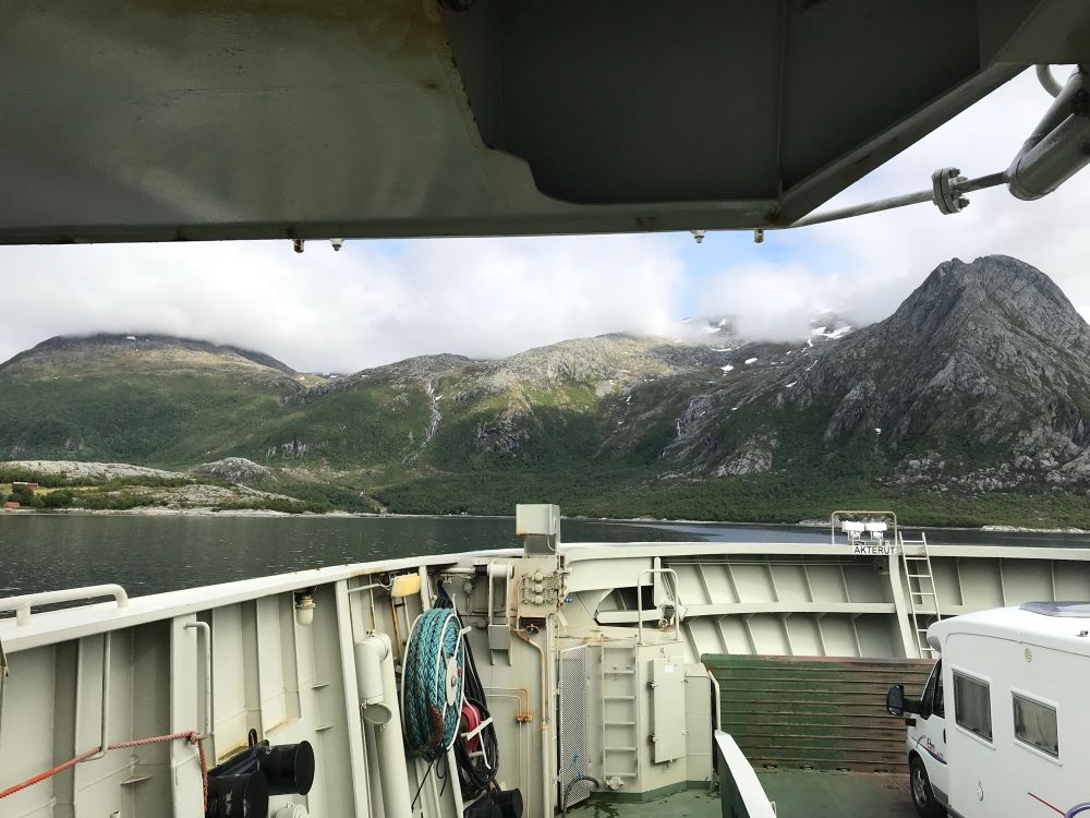 Das Bild zeigt den Blick von einer Autofähre auf eine beeindruckende norwegische Fjordlandschaft. Im Vordergrund sieht man das Deck der Fähre mit einem Wohnmobil auf der rechten Seite und einigen technischen Ausstattungen wie Seilen, Rohren und einem Schlauch auf einer Spule. Die Fähre ist auf einem Gewässer in der Nähe von Vevelstadt unterwegs.

Im Hintergrund ragen steile, felsige Berge auf, die mit grüner Vegetation bedeckt sind. Einige höhere Gipfel sind mit Schneeresten bestückt, und tiefhängende Wolken oder Nebel ziehen über die Bergspitzen hinweg.



