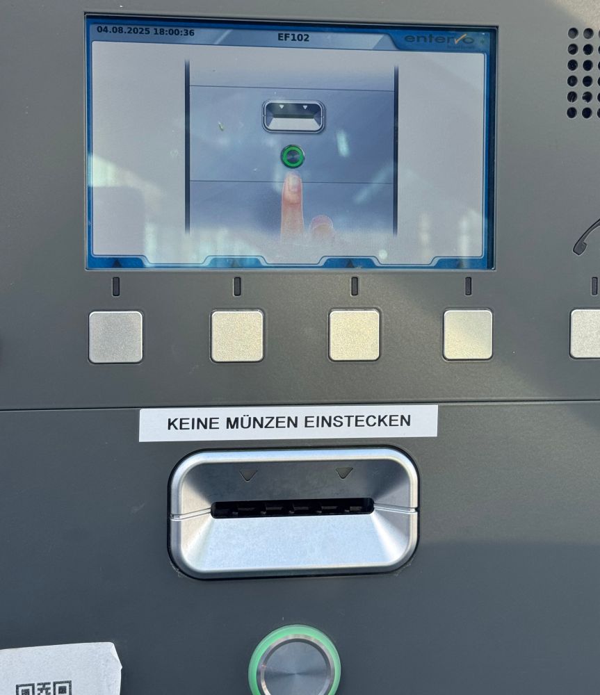 Das Bild zeigt einen modernen Parkscheinautomaten aus Metall. Das Gerät hat ein rechteckiges Display im oberen Bereich, auf dem ein Bild zu sehen ist: Es zeigt einen Finger, der auf einen grünen Knopf drückt – vermutlich eine Anleitung zum Start der Bedienung. 

Unter dem Bildschirm befinden sich fünf quadratische, silberfarbene Tasten, die vermutlich zur Navigation durch das Menü dienen.

Darunter ist ein Kartenschlitz mit der Beschriftung „KEINE MÜNZEN EINSTECKEN“. 

Ganz unten auf dem Automaten befindet sich ein großer runder Knopf mit einem grünen Leuchtring – dieser sieht genauso aus wie der Knopf auf dem Display und ist vermutlich der Start- oder Bestätigungsknopf.
