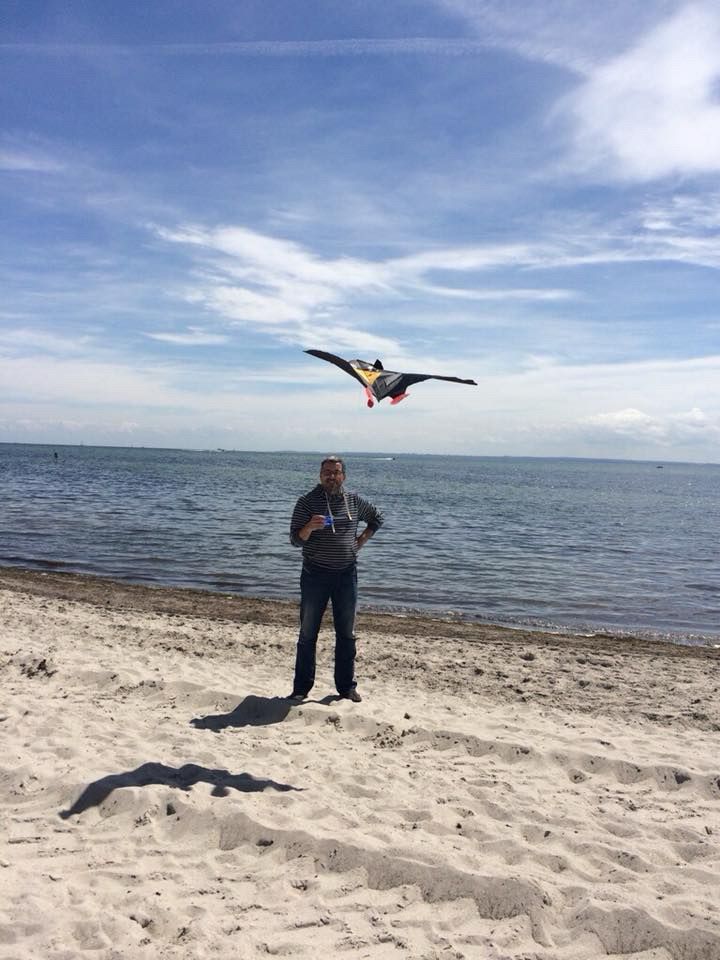 Ein Mann steht an einen Strand, im Hintergrund das Meer. In der Hand hat er einen Drachen, der nur knapp einen Meter über ihm in der Luft schwebt.