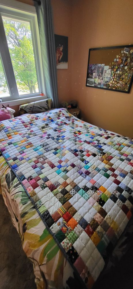 A handmade quilt laid out on a bed. It is primarily white, with many small coloured squares of fabric with various payterns laid out in 4x4 squares