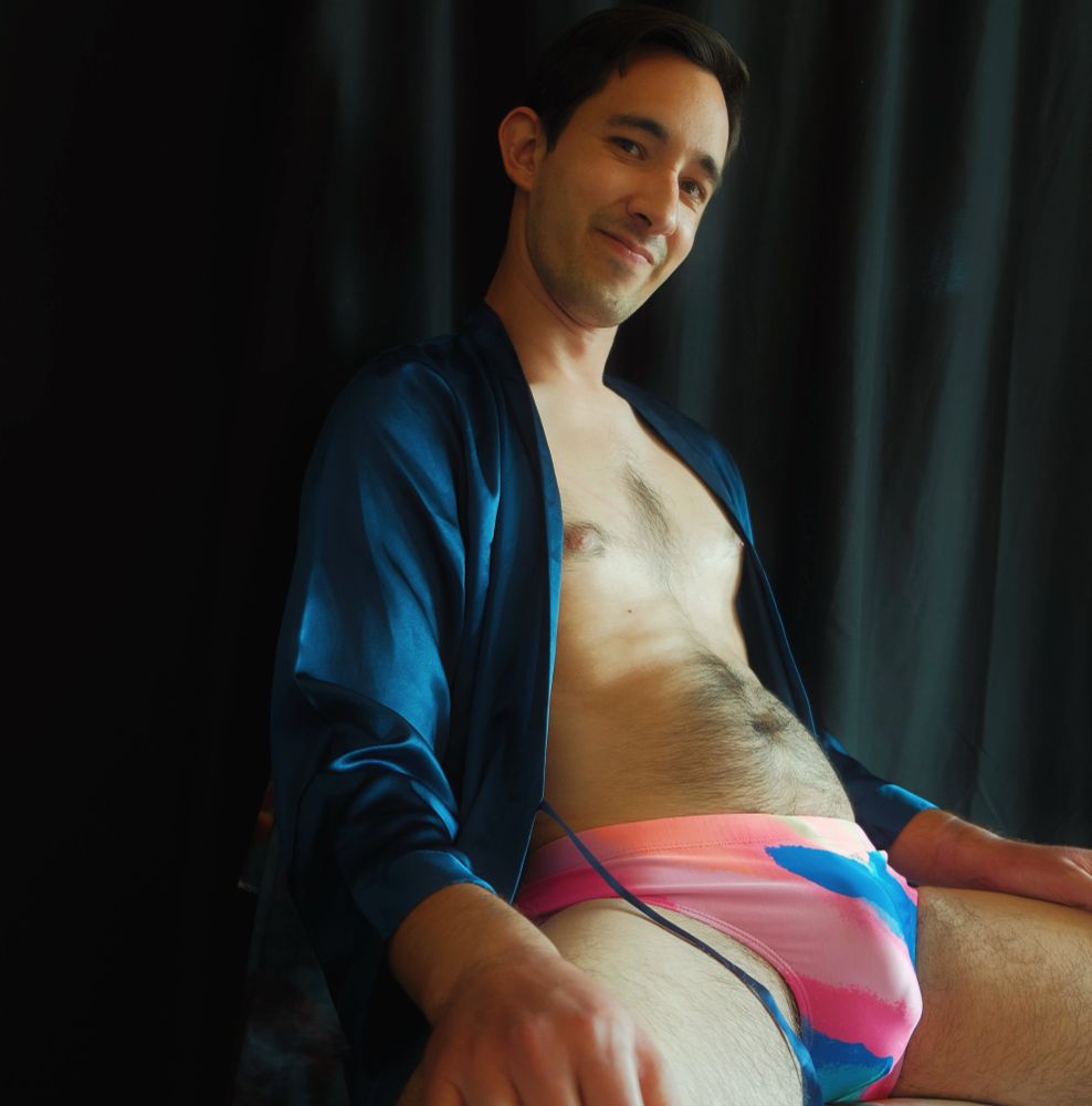 A male model sits in front of a black curtain. He wears an open blue solk robe and pink and blue swim briefs. The briefs accentuate his hard penis and show a bulge. The model cocks his head to his left slightly with a closed-lip smile on his face as he looks into the camera.