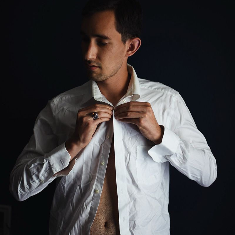 A male model stands against a dark background. He wears a wrinkled white button-down shirt and holds the top button and button-hole in his hands. He looks down and to his right as he attempts to button the shirt. The model also wears a silver ring with a dark orange stone in the center on his right ring finger.