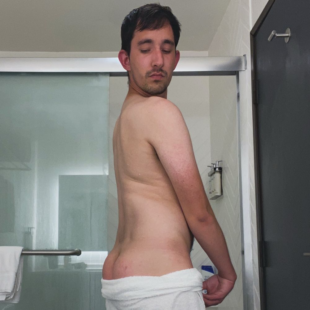 A male model stands in front of a shower with white walls and sliding glass doors. He wears nothing but a white towel, which is slightly below his waist. He shows off the top of his butt while looking down.
