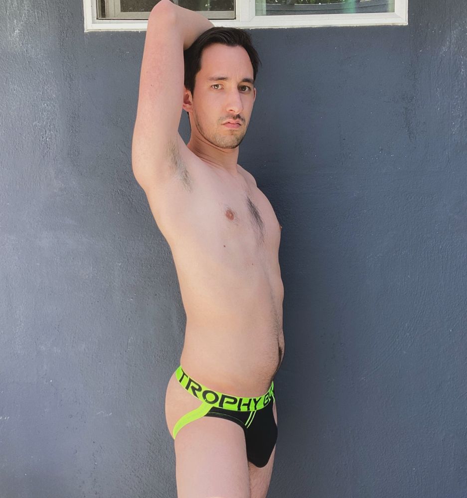 A male model stands against a dark blue wall with a slightly open window. He wears only a black jockstrap with a bright green waistband. The waistband reads “Trophy Boy” in capitalized black lettering. The model rests his right forearm on top of his head, revealing his armpit. His right side is turned toward the camera as he stares sternly.