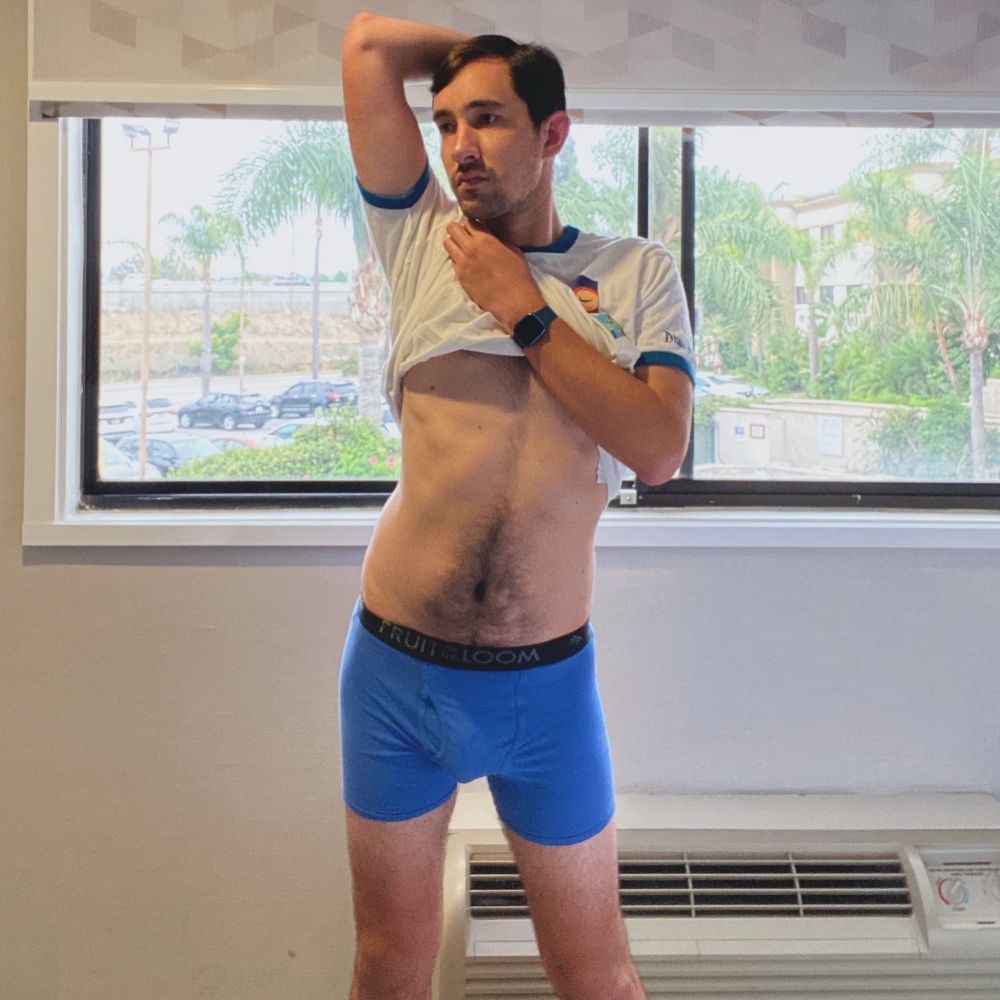 A male model stands in front of a hotel room window and a white wall with a white AC unit. The shades on the window are slightly up, revealing palm trees and a parking lot near a courtyard area. The mode wears a white T-shirt with dark blue cuffs at the sleeve and blue boxer briefs. The model lifts up the shirt with his left hand while his right hand rests behind his head. The model looks off to his right with a blank expression.