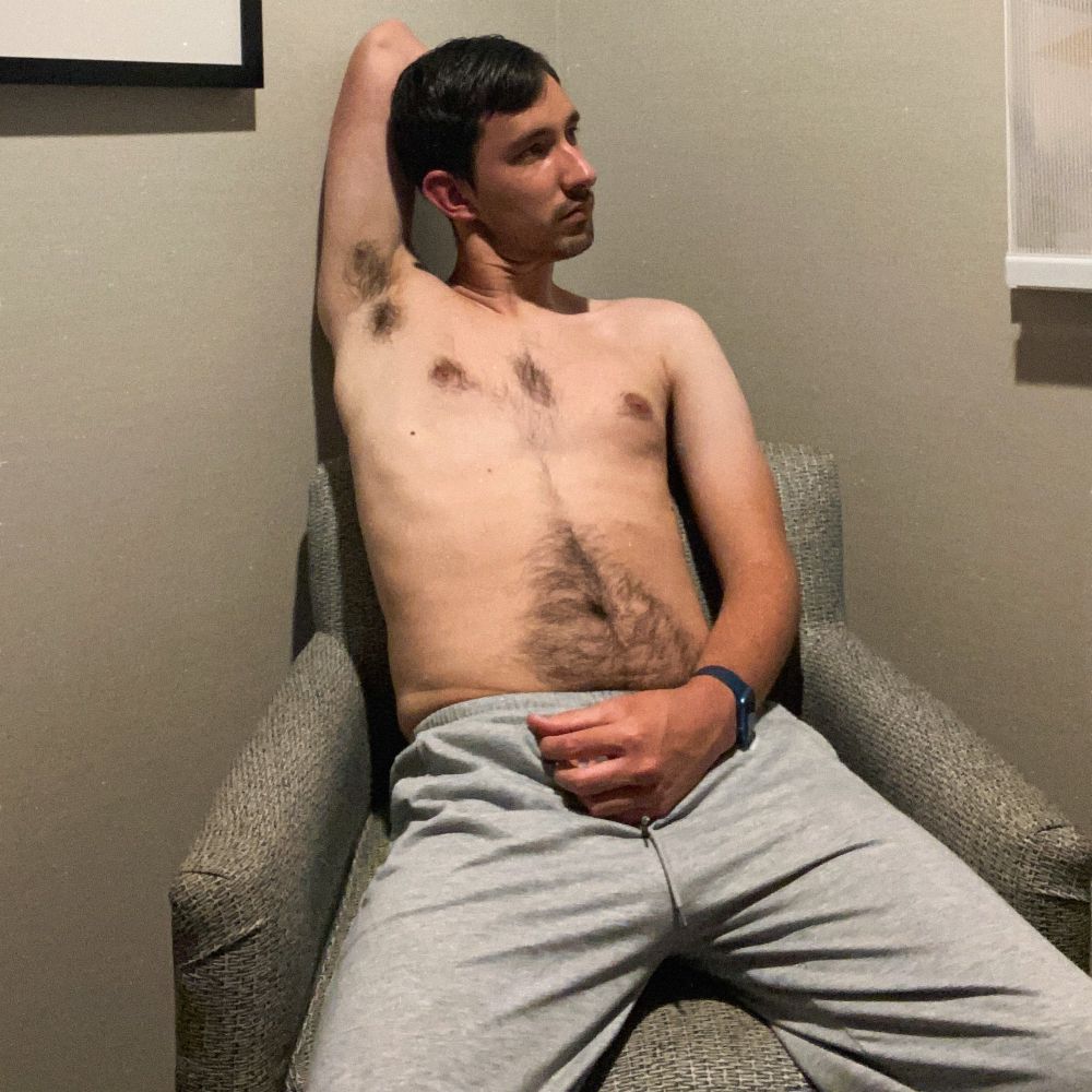 A male model sits on a gray chair in the corner against two white walls. The model wears only gray sweatpants. He puts his right hand behind his head, revealing his armpit. He looks to the left with a blank expression. His left hand rests above the bulge of his sweatpants.