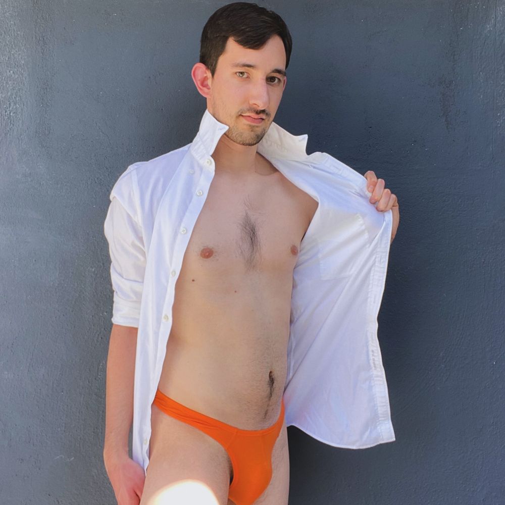 A male model stands against a dark blue wall. He wears an orange swim brief, revealing his bulge. He also wears an open white button-down shirt, which he pulls open with his left hand. He looks into the camera with a slight smirk.