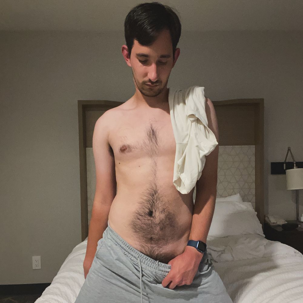 A male model kneels on a bed with white bedsheets and white pillows. He wears gray sweatpants with a white T-shirt on his left shoulder. While looking down, he pulls on the waistband of his sweatpants, slightly revealing his pubes.