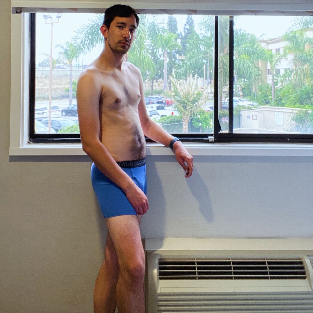 A male model stands in front of a window with white shades drawn up. Behind him are trees and the freeway on a slightly sunny day. The model stands in a pair of blue boxer briefs with his right leg slightly in front. His left forearm rests on the window ledge as he stares into the camera with his head tilted slightly to the left.