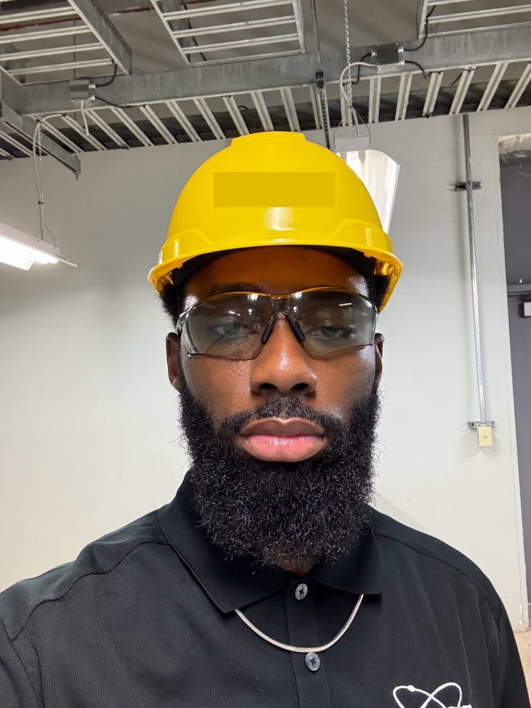 Black man with a beard in a black polo shirt. He has a small necklace on with tinted safety glasses and yellow hard hat. 