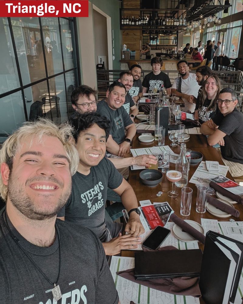 a group selfie of Googlers at the RDU campus eating together and wearing their black Googlers For Job Security tshirts, labeled with “Triangle, NC.”