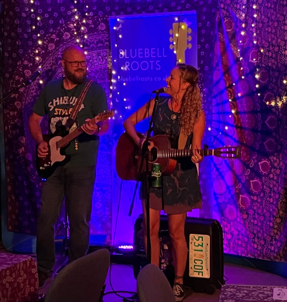 Man plays electric guitar, woman plays acoustic guitar in front of bohemian backdrop with fairy lights and bluebell roots banner 