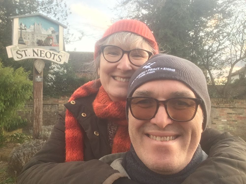 Man and woman smile at camera in front of a decorative town sign for st Neots 
