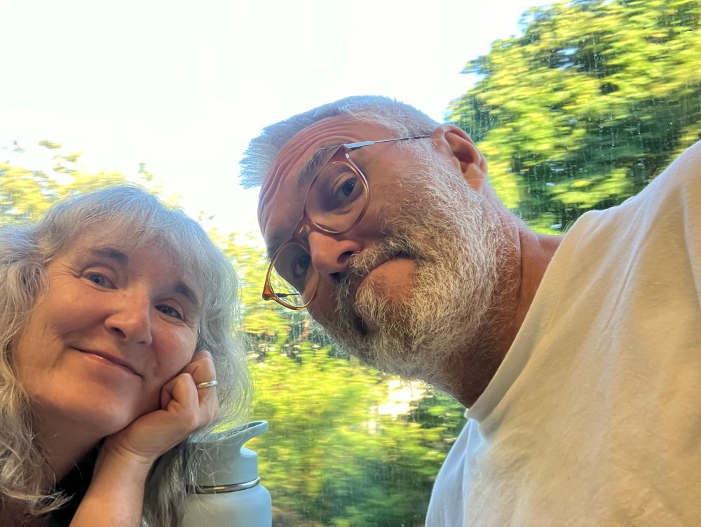 A man and woman smile at the camera in front of a train window rushing past green trees 