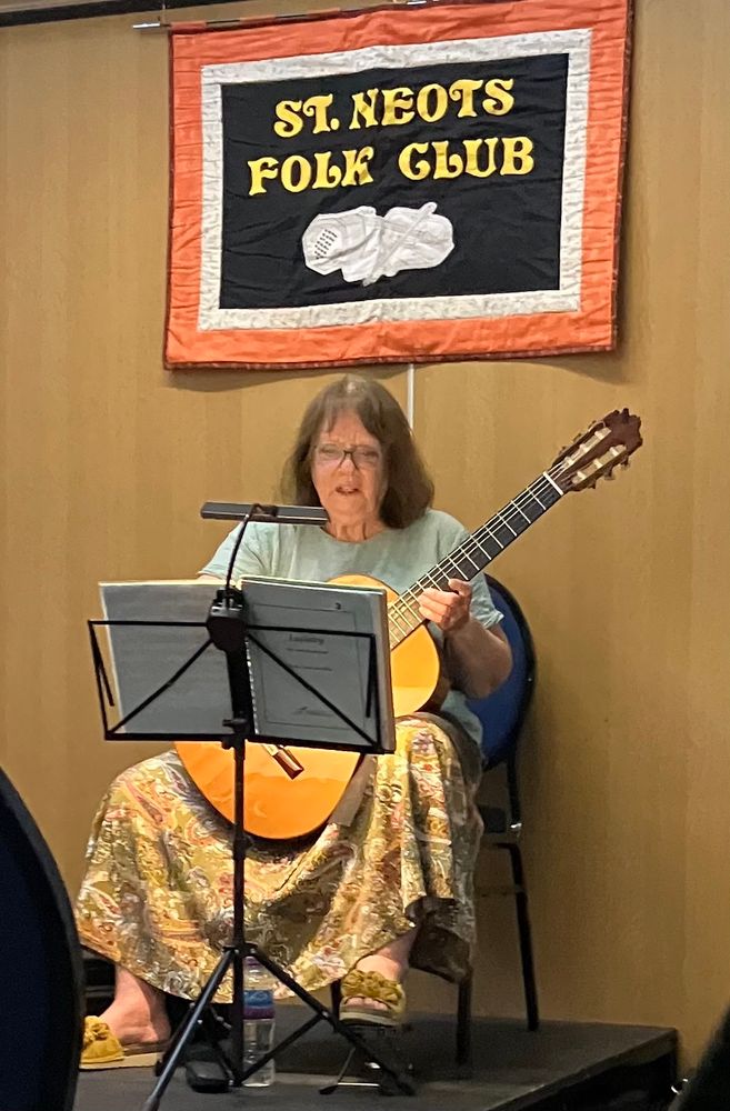 A lady sits playing classical guitar in front of a music stand under a quilted banner saying St Neots Folk Club