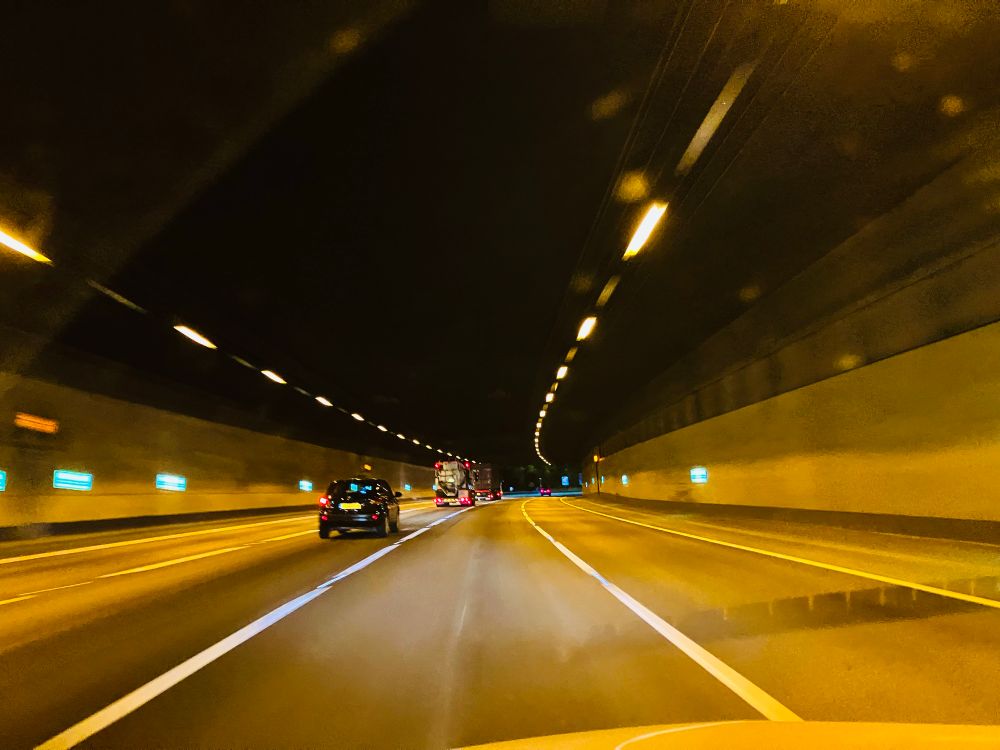 View of road tunnel at night, glowing lights curve into the distance, a car and lorry travel in front 