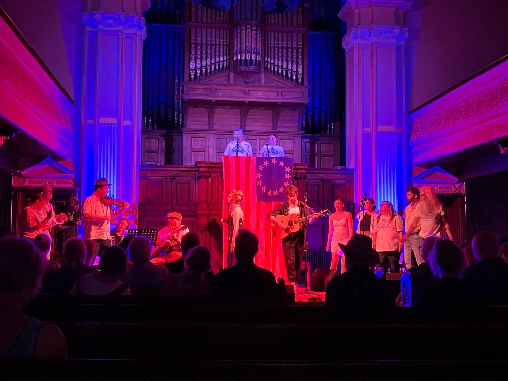 Red and purple lights on stage, audience silhouetted, band and singers at mics, cast to left, chorus to right, two narrators above in front of organ. A finale number is being performed 