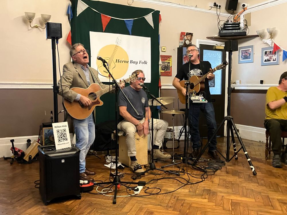 Two men playing guitar one playing cajon in a function room singing into mics