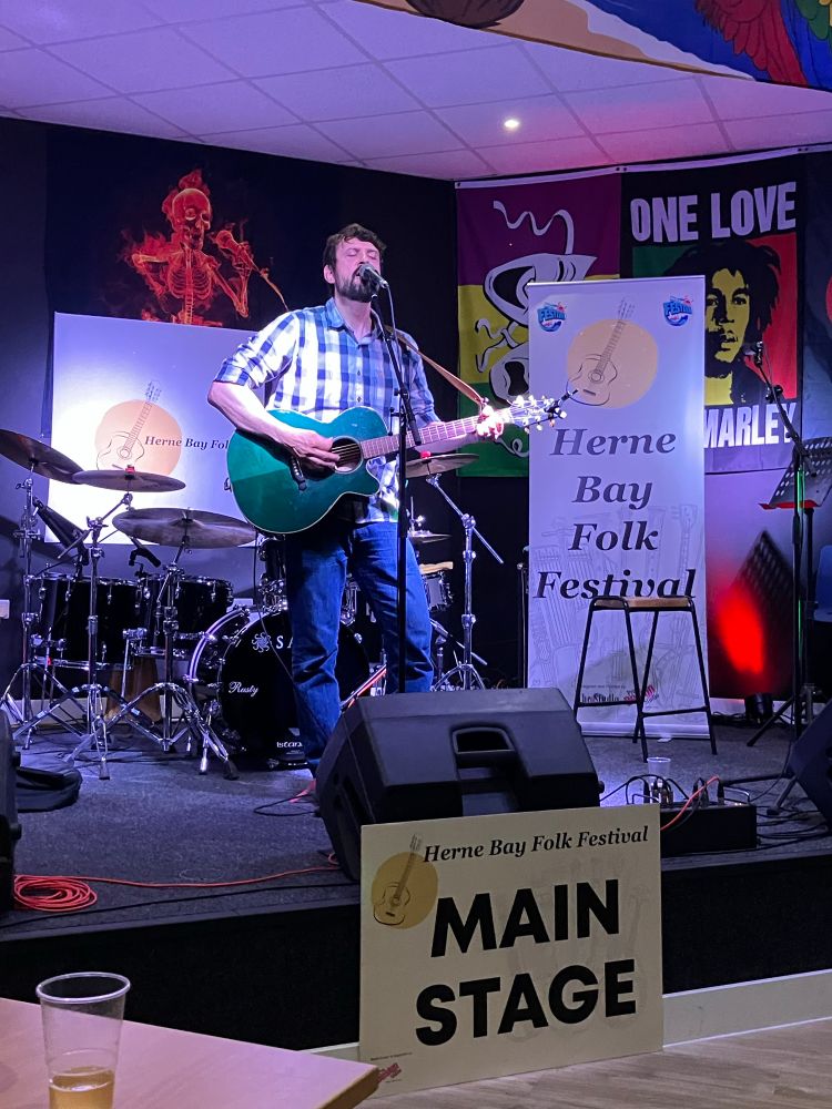 Main in plaid shirt playing guitar singing into mic on stage above sign reading Main Stage