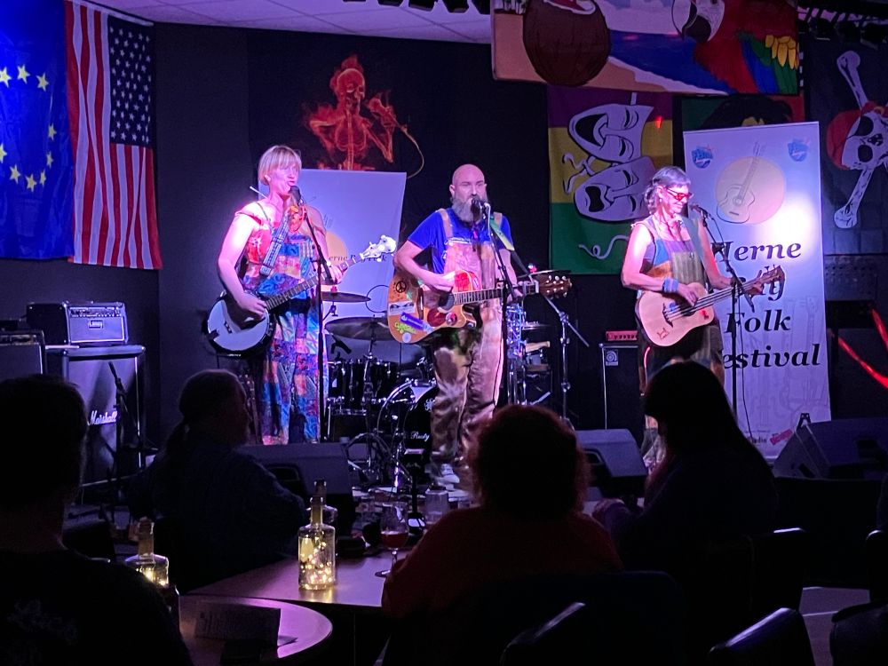 Two woman and a man on stage , 2 play guitar, one plays a banjo, all wear bright colours and dungarees. Audience silhouetted in front of them 
