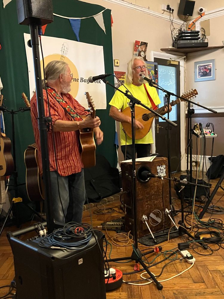 Two men play guitar one sings into a mic, leads and mic stands, speaker and a suitcase drum