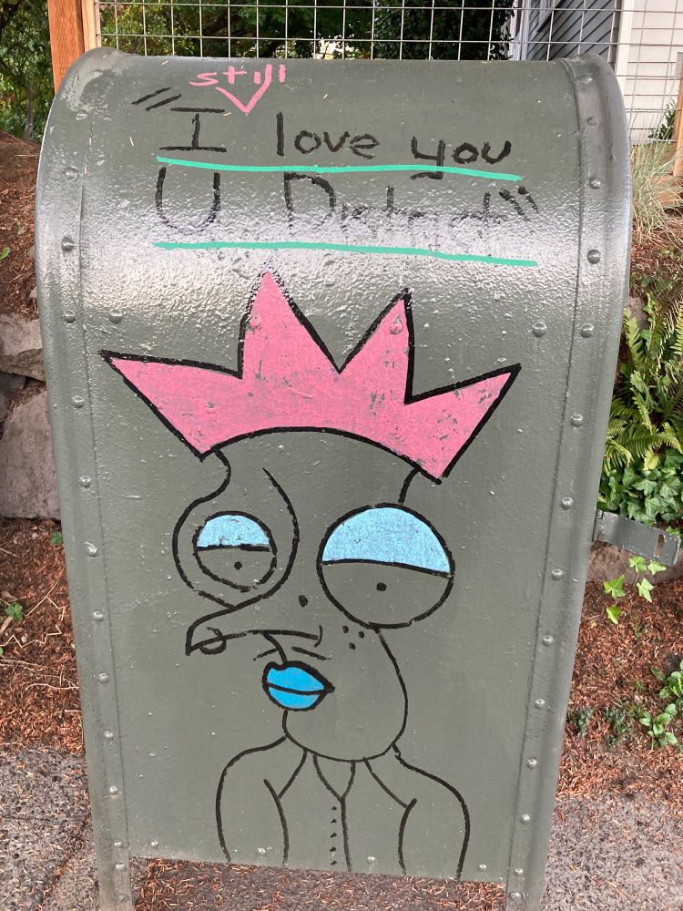 A crowned figured painted on the side of a Seattle mailbox. Its pink crown, blue lips and eyelids pop from the rest of the figure, which is a line drawing. Above the image, a quote "I STILL love you U District"