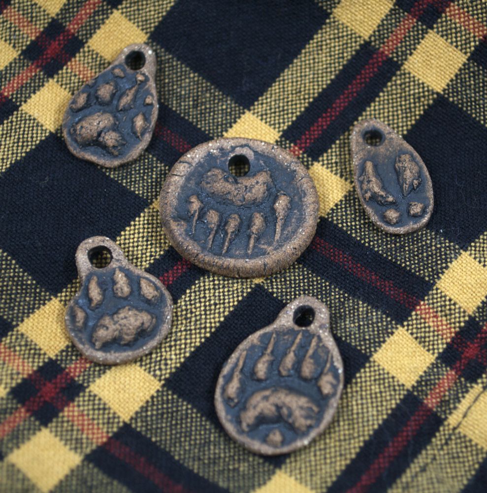 photo showing five stoneware pendants with various animal tracks, sitting on top of a yellow black and red plaid cloth. on the upper left, a canine paw, lower left, a big cat paw, upper middle, a inverted bear paw, lower middle, a bear paw, and upper right, a deer print.