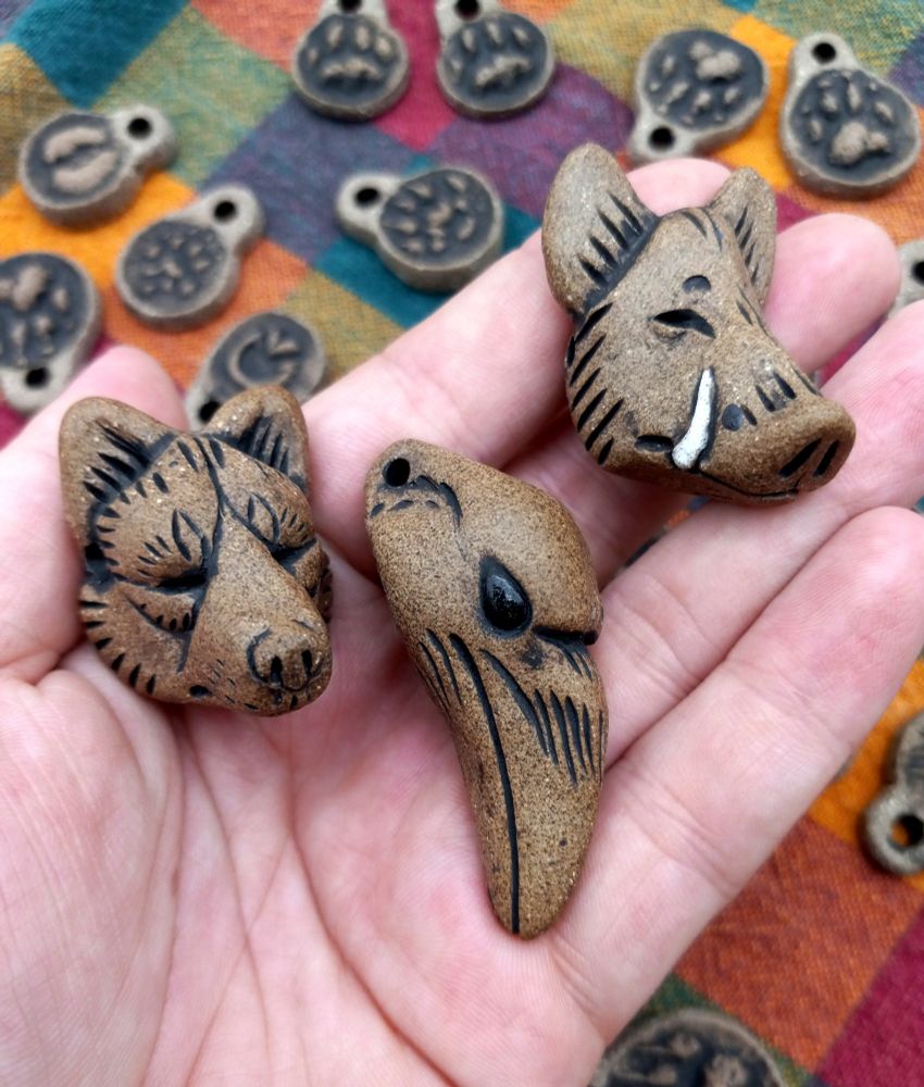 Photo of three animal head shaped clay pendants, the clay is a warm brown that looks almost like stone. They are shaped in the form of a wolf's head, a ravens head, and a boars head. In the background there is colorful plaid fabric with a variety of other animal track pendants scattered on it 