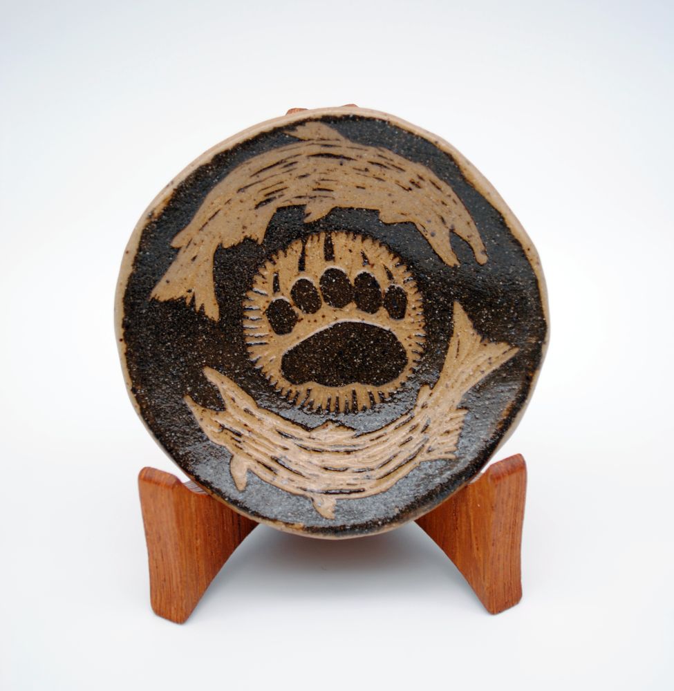 photo of a small ceramic dish sitting on a wooden plate holder. the dish is decorated with carvings depicting a bear paw print and two salmon silhouettes